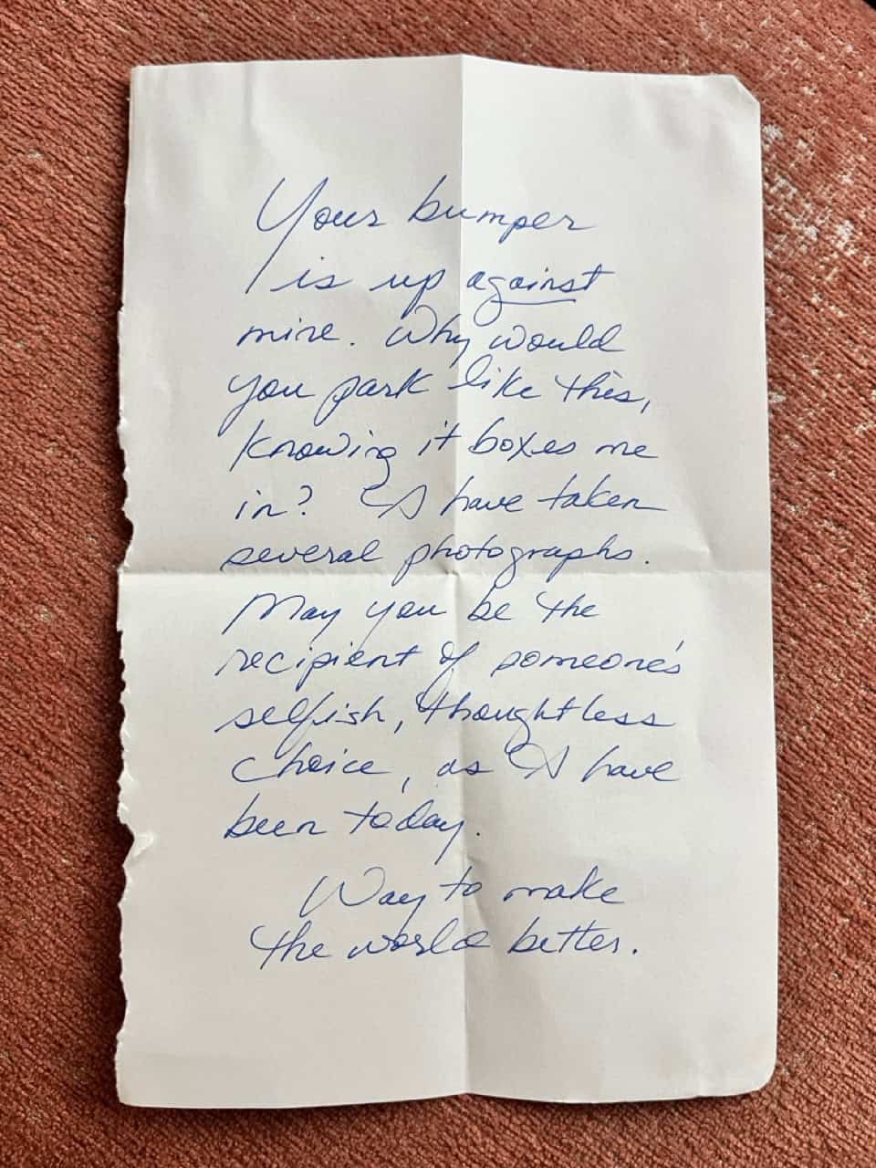 A piece of paper with stylized handwriting in blue ink reads, "Your bumper is up against mine. Why would you park like this, knowing it boxes me in? I have taken several photographs. May you be the recipient of someone's selfish, thoughtless choice, as I have been today. Way to make the world better."