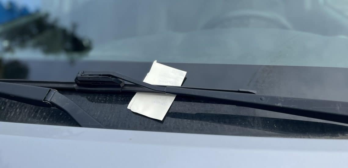 A small rectangular, folded piece of paper pinned under the windshield wiper blade on a silver car, parked and unoccupied.