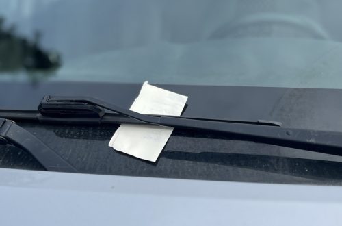 A small rectangular, folded piece of paper pinned under the windshield wiper blade on a silver car, parked and unoccupied.