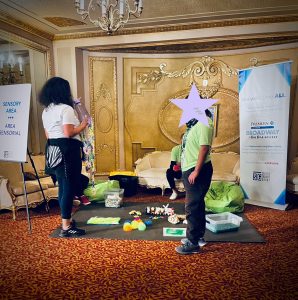 Volunteers in neon-green shirts surround a designated Sensory Area at the northeast end of the First Mezzanine lobby. Vivid colors of the modern toys and sensory items contrast against the background of ornate red and gold brocade of the historic Paramount Theatre's walls and carpeting.